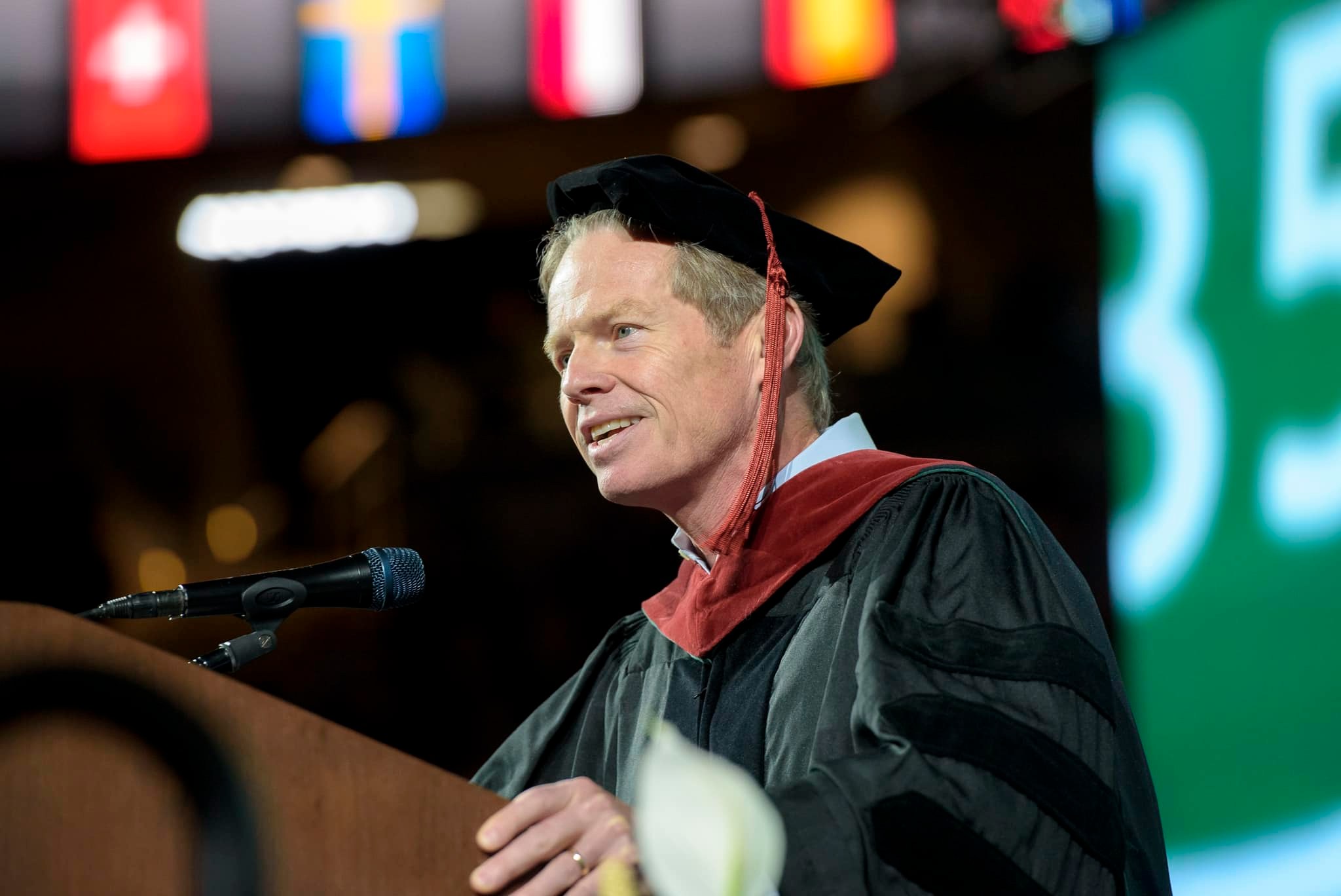 Dr. David Kelly wears his Ph.D. robes and hat while speaking into a microphone at commencement.