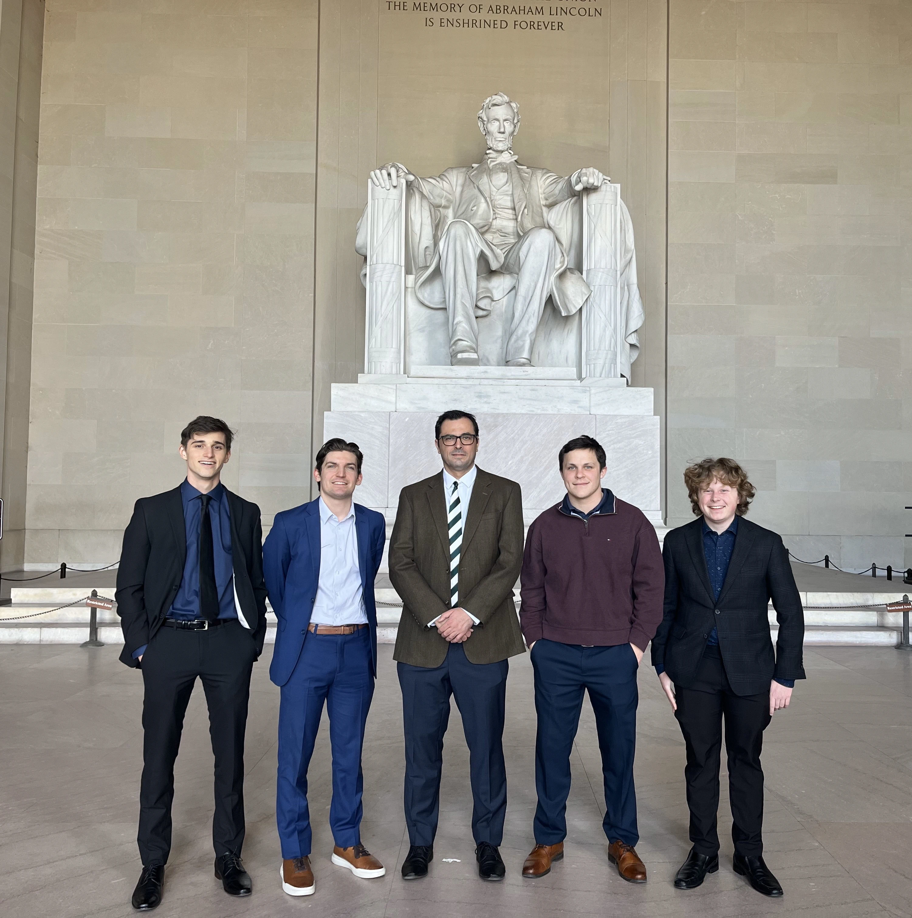 One professor and four students smile in front of the Lincoln Memorial.